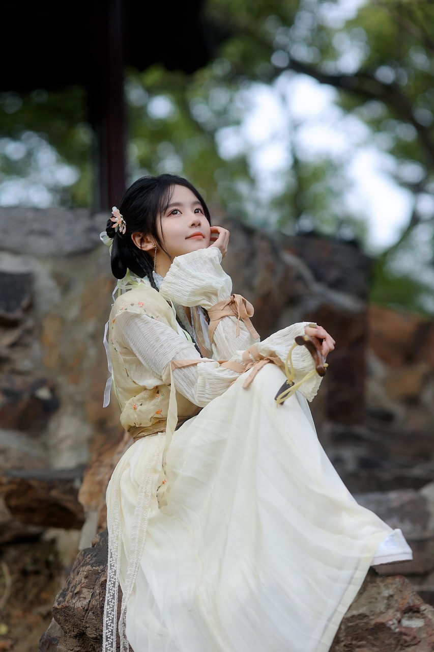 身着浅色汉服的女子坐在石阶上，手持弹弓，望向远方